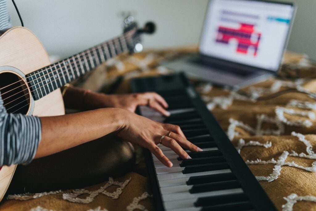 Person holding guitar and playing midi keyboard near computer.